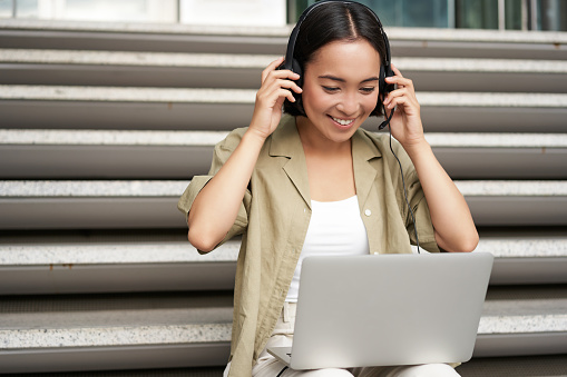 Retrato de una mujer asiática, una niña nómada digital que usa una computadora portátil y escucha música al aire libre. Joven estudiante trabaja en la computadora y sonríe photo