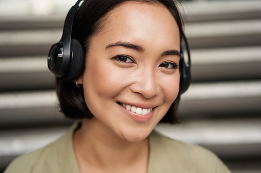 Retrato de primer plano de una chica asiática sonriente con auriculares, escucha música al aire libre, luciendo feliz. photo