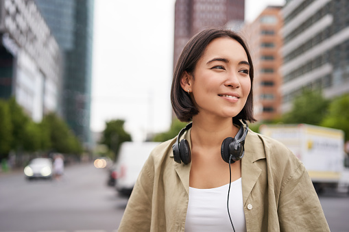 Portrait of young asian woman in headphones, posing in city, smiling and looking away, standing on street of city centre photo