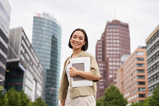 Portrait of young asian woman, looking happy and confident, going to work or university, city skyscrappers behind her, holding laptop and notebook photo