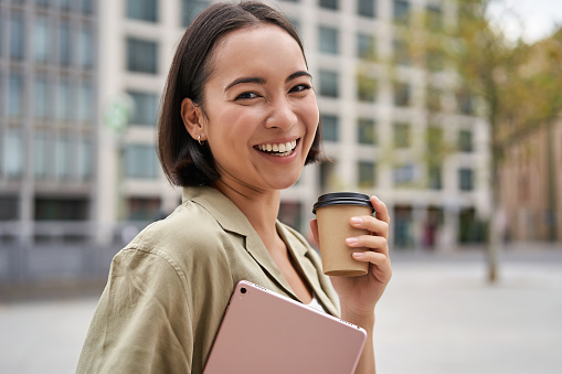 Portrait of asian girl with tablet, drinks coffee on street, walking in city centre photo
