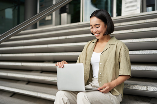 Portrait of young woman student, asian girl using laptop. Asign smiling girl, digital nomad works on her project remotely, sitting on street photo