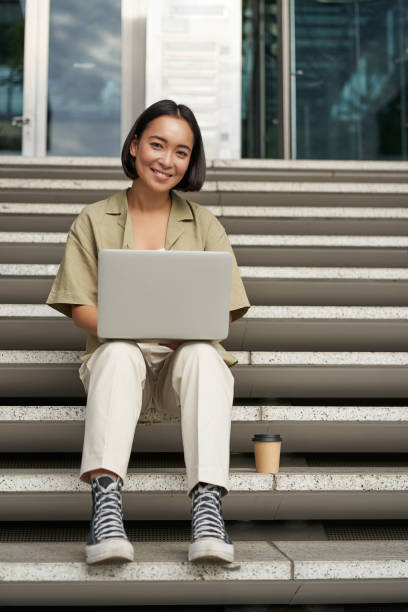istock Young asian woman using laptop outdoors. Cute girl student with computer, does her homework, types, sits on stairs outside campus 2149907282