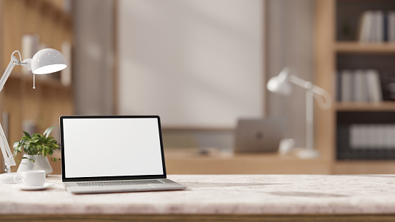 The table showcases a white-screen laptop computer, a coffee cup, a table lamp, and a decor plant in a contemporary office. workspace close-up image. 3d render, 3d illustration