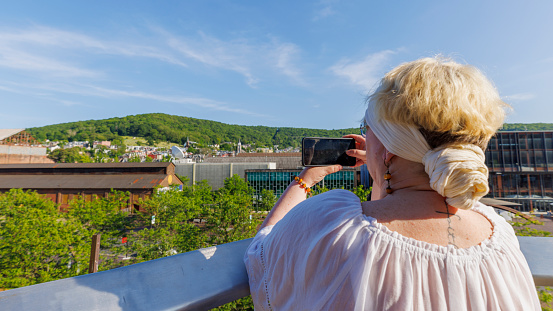 Mature Caucasian blonde woman with tattoo, visiting Steelstacks in Bethlehem, Pennsylvania. She captures the vibrant, green landscape and industrial remnants using her smartphone in a sunny day