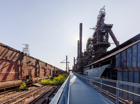 Historic Steelstacks, an abandoned steel factory in Bethlehem, Pennsylvania, featuring a long walkway leading towards massive old rusted structures, set against a clear sky. The empty tracks and aged buildings reflect the industrial past and resonate with themes of industrial decline and historical preservation.