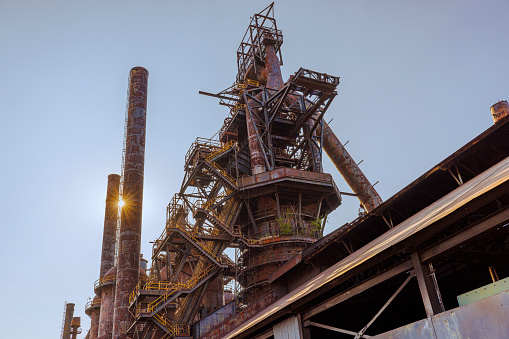 Sunlight beams through the rusty structure of the Steelstacks, an abandoned steel factory in Bethlehem, Pennsylvania. The intricate ironwork and the grand scale of the industrial age, now silent and still, set against a clear sky at sunset