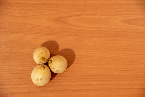 Three langsat fruit on a wooden surface, one with a natural marking resembling a face. These tropical fruits are edible, with yellow skin and juicy, sweet, and tangy flesh. They exude an aromatic fragrance and are rich in vitamin C and dietary fiber. A healthy snack, they offer antioxidants and are a rare find in Southeast Asian cuisine. Copy space for text