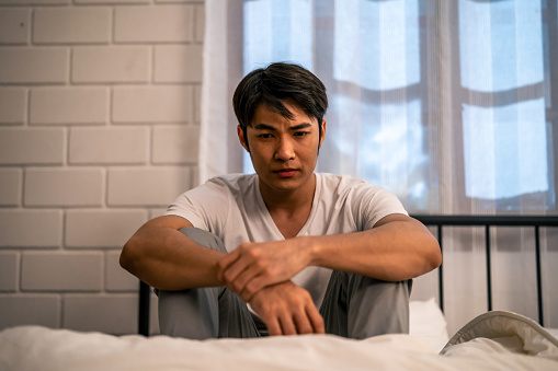Asian frustrated insomnia man in pajamas sit on bed in dark night time. Attractive depressed young sleepless man feeling upset, worried and concerned cannot sleep from nightmare in bedroom in home.