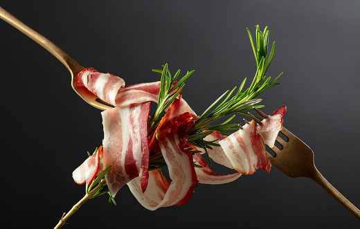 Dry-cured pork belly bacon with rosemary on a black background. Sliced bacon on a forks.
