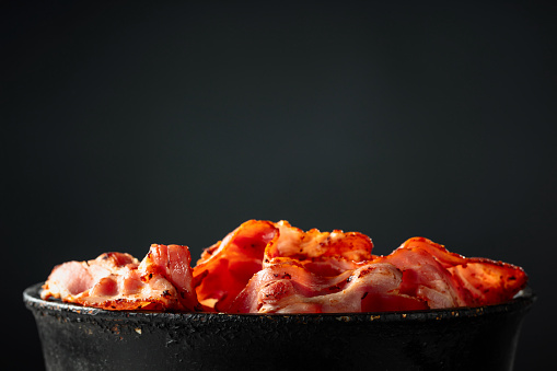 Fried bacon slices in old black pan on a black background. Copy space.