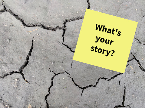 A yellow sticky note with the intriguing question Whats your story? written in bold black ink. The note is placed against a stark, cracked gray surface, evoking a sense of stories and experiences emerging from fractured beginnings.