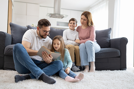 A family of four enjoys a cosy moment with a book at home, showcasing the bond between parents and kids Keywords: mother, kid, cosy