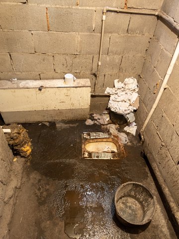 Dirty toilet room with genoa bowl, pile of used toilet paper and a bucket, uncoated concrete flooring and cinder brick walls. Developing country poorness.