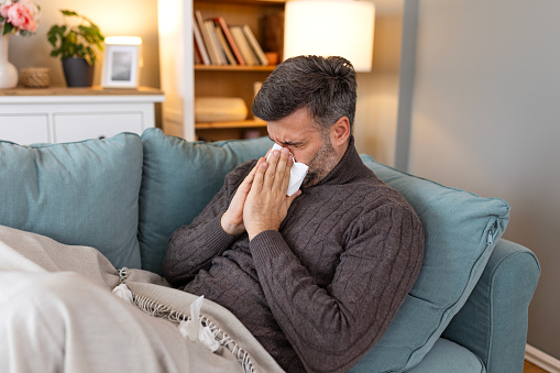 Sick sad man sits on couch at home suffers from runny nose flu disease coronavirus pandemic covid epidemic sneezes. Unwell guy feeling bad fever virus illness symptoms indoor