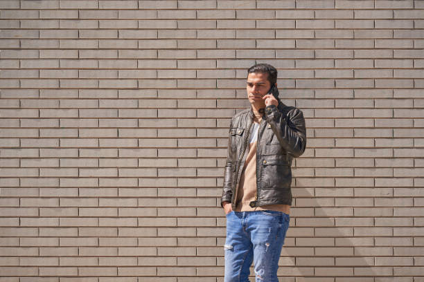 Young man wearing black jeans, a black leather jacket and sunglasses sitting in front of a brick wall looking at the camera