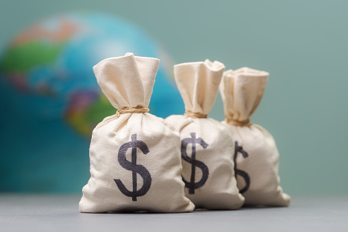 Three money bags with dollar signs in the foreground, and a blurred globe in the background, representing global finance