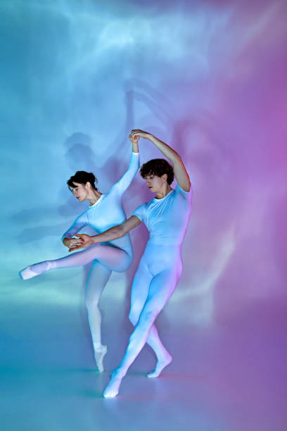 Two elegant ballet dancers in synchronous pose, female dancer holds one leg raised, against blue and purple shadowy background. Concept of beauty and elegance, dance grace, inspiration, creativity.