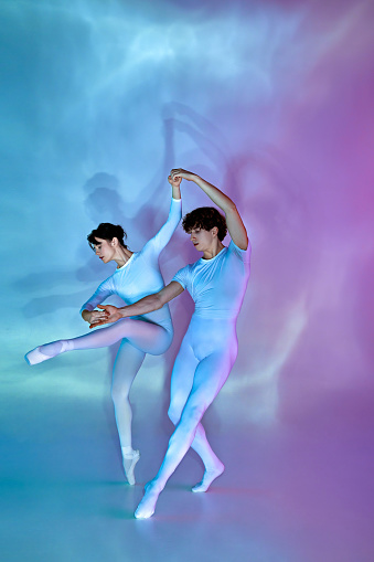 Two elegant ballet dancers in synchronous pose, female dancer holds one leg raised, against blue and purple shadowy background. Concept of beauty and elegance, dance grace, inspiration, creativity.