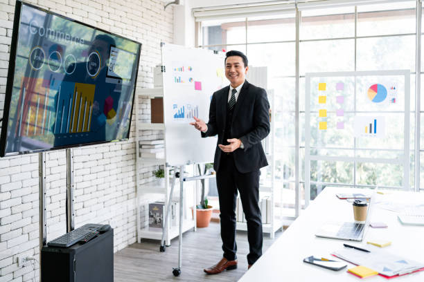 istock A cheerful and confident Asian businessman stands, presenting bar charts data from the projector screen to his office colleagues. Asian businessman leader role at the meeting. 2149930599