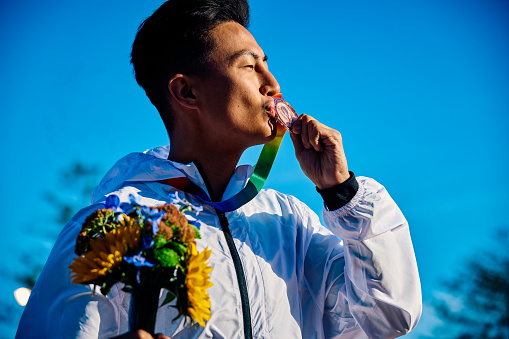 International athlete kissing medal with bouquet of flowers. Outdoor portrait with architectural backdrop. Reflection on success concept