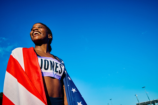 Smiling athlete wrapped in American flag. Outdoor portrait with blue sky. Patriotism and national pride concept.