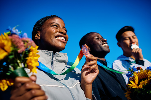 Group of International athletes with medals and bouquets celebrating victory. Team success outdoor portrait under blue sky. Multicultural unity and sportsmanship concept.