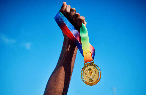 raised hand holding a gold medal against blue sky. victory and achievement concept. - vencer imagens e fotografias de stock