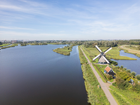 The calmness of the Dutch countryside is quietly reflected in this wide-angle view. A traditional windmill behind a brown roofed bungalow located by a calm canal, displays its sails in a gentle pause. The water's surface mirrors the scene, adding to the overall peacefulness. On the side, a narrow motorable path is seen. The clear blue sky overhead and the lush greenery along the canal’s edge enhance the simple beauty of the scene.