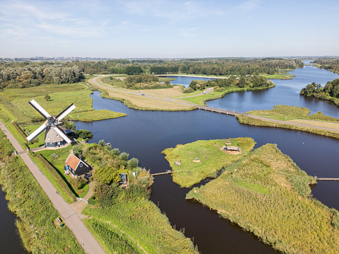 Aerial view of a typical Dutch scene with a windmill at the crossroads of winding canals. On the side, a narrow motorable path is seen and in the distance, the green pieces of land surrounded by the calm water. The area is open and not crowded, giving a feeling of living quietly alongside nature.