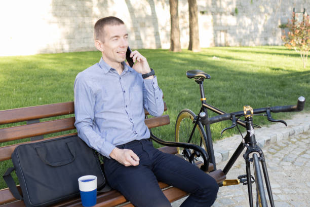istock Smiling businessman commuter using mobile phone. He is enjoying at public park on his bicycle trip from work after a long workday. 2149939327