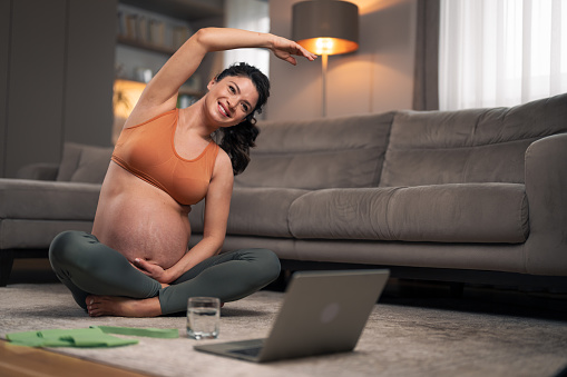 A beautiful and healthy pregnant lady doing yoga stretching exercises while following a video tutorial on her laptop in the living room.