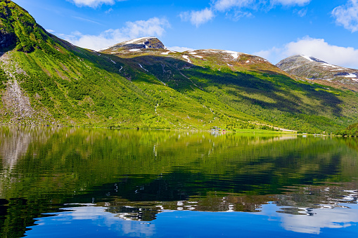 Green reflections in cold water. Lake Eidsvatnet. The water reflects lush clouds, blue skies and forested shores. Picturesque mountains in central Norway. Summer in Scandinavia.