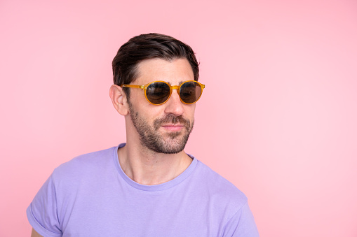 Portrait of a stylish man in purple t-shirt and trendy sunglasses posing with a confident smile on a soft pink backdrop
