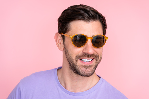 Handsome young man wearing yellow sunglasses posing with a confident smile against a vibrant pink background, showing casual summer vibes