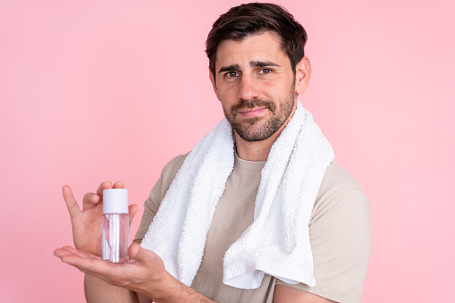 Young adult male with towel around neck looking puzzled while holding a bottle of skincare lotion on a pink background
