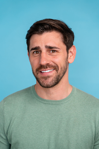 Portrait of a smiling young man with dark hair and stubble, wearing a green sweater against a blue background