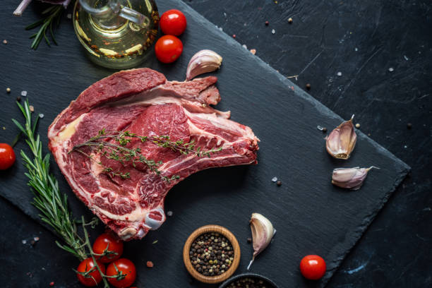 istock Raw beef steak on the bone on a black slate serving plate, decorated with various ingredients such as cooking oil, dry pepper, cherry tomatoes and herbs. 2149957024