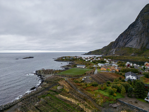 Gray skies hang over a quiet seaside village next to Norway's towering cliffs. The community is clearly seen in groups of houses with red and white tops, painting a picture of tight bonds among neighbors. Nearby, wooden racks used for drying fish speak to a life linked with the sea. The steps of farmland etched into the cliffs show the cleverness of the locals in using their land well. A soccer field adds a current spin to the village's leisure, as the open sea and sky stretch out.