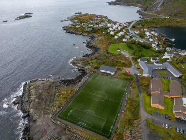 Gray skies hang over a quiet seaside village next to Norway's towering cliffs. The community is clearly seen in groups of houses with red and white tops, painting a picture of tight bonds among neighbors. Nearby, wooden racks used for drying fish speak to a life linked with the sea. The steps of farmland etched into the cliffs show the cleverness of the locals in using their land well. A soccer field adds a current spin to the village's leisure, as the open sea and sky stretch out.