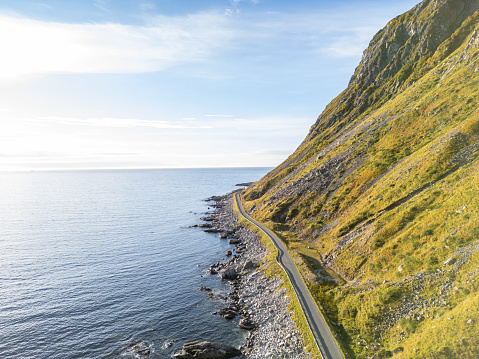 A winding road following a rough coastline. The warm light from either sunset or sunrise illuminates the rocky shore and sparkling sea. The road, fitting closely to the land's shape, provides stunning views of the sea and horizon, promising adventure. With no cars or people visible, the scene feels peaceful and undisturbed.