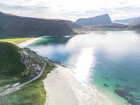 A peaceful scene where land, sea, and light meet at a beautiful fjord. A winding road runs along green fields that become a sandy, curved beach. Sunlight reflects on the water, making it sparkle and showing the different depths and the seabed. Beyond the beach, tall mountains rise up, forming a striking and beautiful skyline under the soft light of the sky.