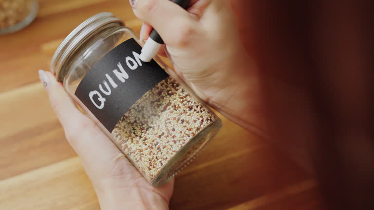 Close-up  hands writing 'Quinoa ’ with black label on a glass jar filled  on a wooden counter for organized kitchen pantry storage ,zero waste Lifestyle with green shopping,Plastic free and sustainability at home