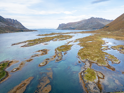 A mosaic of shallow waters and scattered islets that unfolds into a valley embraced by towering mountains, their slopes adorned with the muted hues of vegetation greeting the cooler season. The placid water mirrors the vast sky, enhancing the tranquility and breadth of the Nordic landscape. This image invites viewers into a realm of peace, spaciousness, and the beauty of the north's untouched wilderness.