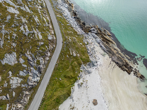 Seen from above, is the graceful curve of a coastal road winding along the boundary of a rocky shoreline and a white sandy beach. The vivid blue sea contrasts sharply with the pale sands and the soft colors of the grass and rocks beside the road. The natural shapes of the landscape and the designed road come together in a peaceful mix of nature and human construction, bringing out feelings of calm and the charm of seaside travels.