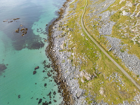From an aerial viewpoint is a single coastal road in between small green plants that winds along a bay with transparent turquoise waters. The shoreline, dotted with stones and boulders, stands in stark contrast to the calm clear sea.