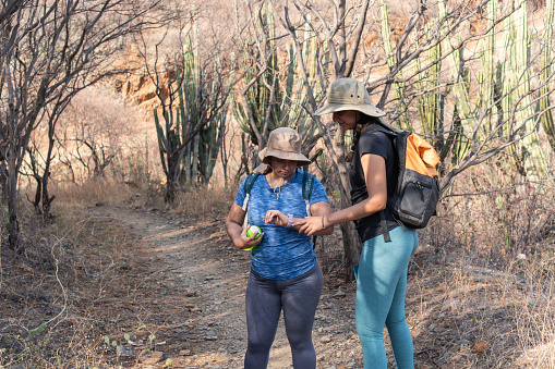 Two women pause their hike to look at a smartwatch together on a dry, forested trail with cacti in the background.
