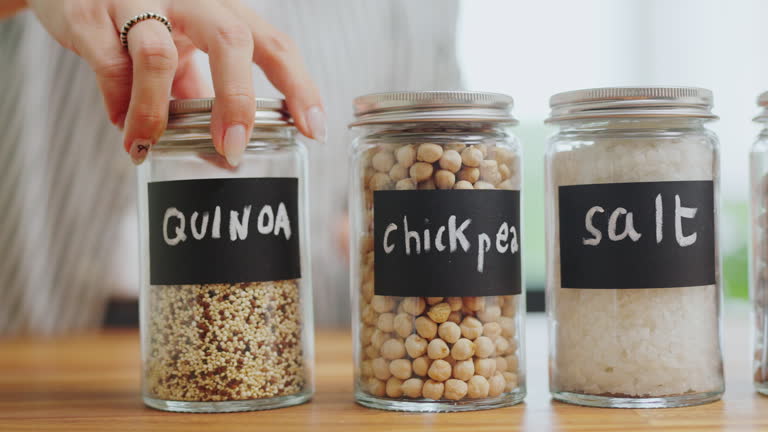 Close-up  hands writing 'Quinoa ’ with black label on a glass jar filled  on a wooden counter for organized kitchen pantry storage ,zero waste Lifestyle with green shopping,Plastic free and sustainability at home
