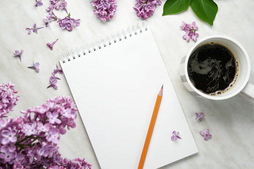 Spring lilac flowers on workdesk female home office with notebook, pensil and coffee white background. Top view, flat lay, mockup.