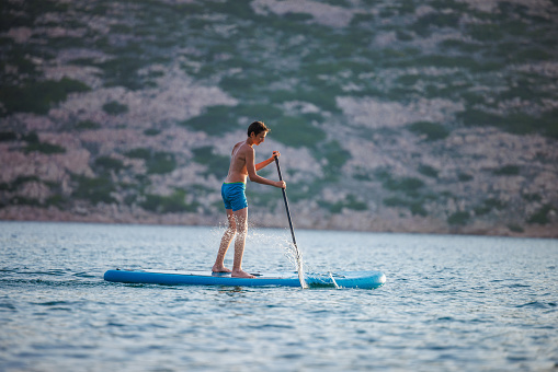 Teenage boy in blue trunks paddle boarding on water surface and rowing with a splash, active summer vacation at the seaside
